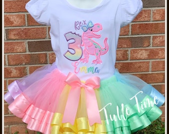 Può includere: Una maglietta bianca con un dinosauro rosa con un fiocco sulla testa e il testo "Rex" ed "Emma" ricamato su di essa. La maglietta è abbinata a una gonna di tulle arcobaleno con più strati di nastro di raso.