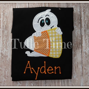 May include: Black t-shirt featuring a white ghost holding a candy corn. The candy corn has orange polka dots and yellow gingham patterns. The name "Ayden" is embroidered in orange thread below the graphic. The shirt is a Halloween-themed design.