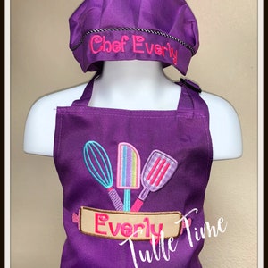 May include: Purple chef hat and apron set with the name "Chef Everly" embroidered on the hat and "Everly" embroidered on the apron with a banner and three kitchen utensils. The apron has a pocket and the text "Tulle Time" is visible in the bottom right corner.