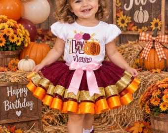 May include: A white short-sleeve shirt with a pumpkin and floral graphic featuring the text Two and Vienna, paired with a layered burgundy, gold, and orange tutu skirt with a pink satin bow, and white ruffled socks with burgundy bows.