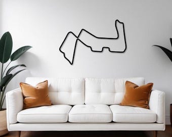 May include: Black metal wall art depicting a race track outline. The artwork is mounted on a white wall above a white sofa with two orange-brown throw pillows. Green plants in woven baskets are on either side of the sofa.