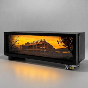 Puede incluir: Una caja de luz rectangular negra con una escena iluminada en tonos naranja del edificio Energetik en Prípiat. La pantalla muestra la estructura con texto cirílico en el techo, árboles silueteados y un fondo de atardecer brillante.