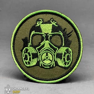 May include: Circular embroidered patch with a neon green gas mask design on an olive green backdrop. The patch has a black border and the text "CHORNOBYL SHOP" at the bottom. The gas mask design is detailed and prominent.