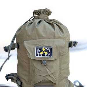 May include: Olive green drawstring backpack with a front pocket and adjustable straps. A patch on the pocket displays a yellow and black hazard symbol with the text "CHORNOBYL.TOUR.COM". The bag has a button closure and a cinched top.