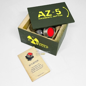 Könnte beinhalten: Ein roter runder Knopf mit Metallrand in einer grünen Holzkiste. Der Deckel trägt den gelben Schriftzug AZ-5 CHORNOBYL BUTTON und ein Strahlensymbol. Eine beiliegende beige Karte zeigt denselben Knopf mit einem beschreibenden Text.