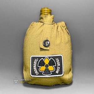 May include: A khaki-colored canteen with a matching fabric cover. The cover has a black button closure and a patch with a yellow and black hazard symbol and the text "CHORNOBYL TOUR.COM". The canteen's cap is a golden color.