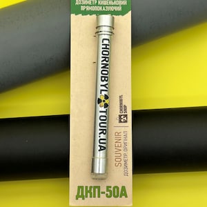 May include: A silver pocket dosimeter with the text "CHORNOBYL TOUR.UA" and "SOUVENIR" printed on it. The dosimeter is on a green and white card with Ukrainian text and the words "Pocket Direct Reading Dosimeter". The background is yellow and black.