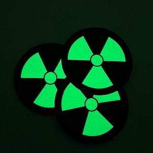Könnte beinhalten: Drei schwarze, kreisförmige Untersetzer mit einem leuchtend grünen Strahlungssymbol. Die Symbole leuchten vor dem dunklen Hintergrund. Die Untersetzer sind leicht überlappend angeordnet.