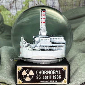 Könnte beinhalten: Eine Schneekugel mit einem detaillierten Modell des Kernkraftwerks Tschernobyl. Das weiße und rote Modell befindet sich in einer transparenten Glaskugel. Auf dem Sockel steht "CHORNOBYL 26 april 1986".