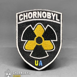 Pode incluir: Patch bordado em forma de escudo com a palavra "CHORNOBYL" acima de um símbolo de radiação amarelo e preto. As letras "UA" em azul estão na parte inferior. O patch é da loja "CHORNOBYL SHOP".