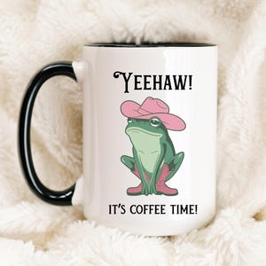 May include: White coffee mug with a black rim and handle. The mug features a cartoon frog wearing a pink cowboy hat and boots. The text "Yeehaw! It's coffee time!" is printed on the mug.