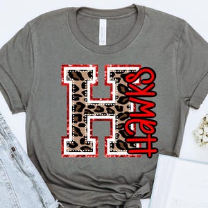 May include: A gray t-shirt with a red and black leopard print letter H and the word "Hawks" in red.