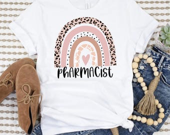 May include: White t-shirt with a rainbow design featuring a pink, brown, and white color scheme. The rainbow has a leopard print pattern and a small pink heart in the center. The text "Pharmacist" is printed below the rainbow.