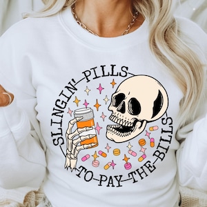 May include: White sweatshirt with a graphic of a skull holding a pill bottle and the text "Slingin' Pills to Pay the Bills".