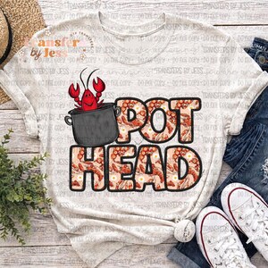 May include: A light-colored t-shirt with the words "POT HEAD" in a bold, outlined font. The letters are filled with a crawfish pattern. A cartoon crawfish is in a pot above the word "POT".