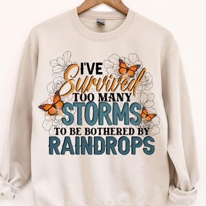 May include: A beige sweatshirt with a graphic print of butterflies and flowers. The text on the shirt reads "I've Survived Too Many Storms To Be Bothered By Raindrops".
