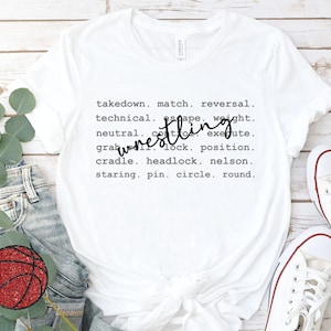 May include: White t-shirt with the word "wrestling" in a cursive font. The shirt features a list of wrestling terms in a simple, sans-serif font, including "takedown," "match," and "reversal."
