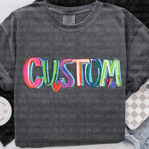 May include: Dark gray long-sleeve shirt with the word "CUSTOM" in colorful bubble letters. The word has a heart design. The shirt is displayed with a phone case and jeans.