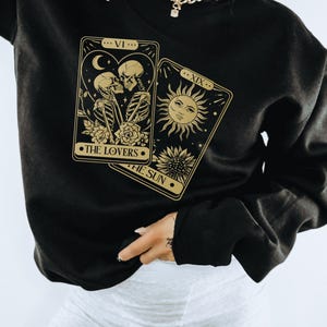 May include: Black sweatshirt with a gold tarot card design featuring The Lovers and The Sun cards. The cards are illustrated with skeletons and a sun.