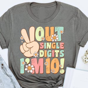 May include: A gray t-shirt with a colorful graphic that says "Out Single Digits I'm 10!" with peace sign and flowers.