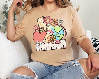 May include: A tan t-shirt with a graphic design of a red apple, a pencil, a globe, and a ruler. The graphic is surrounded by pink and white flowers.