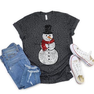 May include: A dark gray t-shirt with a leopard print pattern featuring a sequined snowman design. The snowman has a black top hat, a red scarf, and a carrot nose. The image also includes blue jeans and white sneakers.