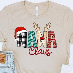 May include: Beige t-shirt with a Christmas themed design that says "Nana Claus". The design features a Santa hat, reindeer antlers, and plaid patterns.