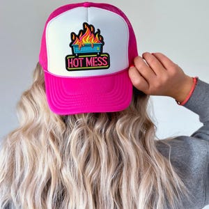 May include: A pink trucker hat with a white front panel and a mesh back. The hat features a colorful graphic of flames above a blue container with the words "HOT MESS" in a neon-style font. The hat is worn by a person with blonde hair.