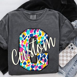 May include: A gray sweatshirt with a large, colorful letter "C" featuring a custom design. The letter is filled with a vibrant pattern of blue, pink, yellow, and white shapes. The word "Custom" is written in a cursive font across the letter. A phone with a checkered case rests nearby.