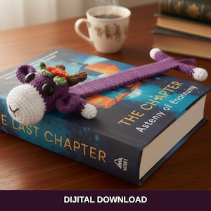 Puede incluir: Un marcapáginas de ganchillo hecho a mano con forma de reno morado, apoyado en un libro titulado "The Chapter". El reno tiene una cara blanca, ojos negros y cuernos marrones con detalles en naranja y verde. El marcapáginas está sobre un libro con una cubierta azul. Una taza de café está cerca.