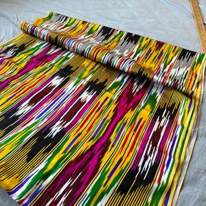 May include: A colourful fabric with a striped pattern in shades of yellow, black, green, pink, blue, and white. The fabric has a traditional ikat design.