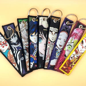 May include: A collection of embroidered keychains featuring anime character designs. Each keychain has a rectangular shape with a metal ring and depicts a different character in vibrant colors. The keychains are arranged in a fan shape against a light yellow background.
