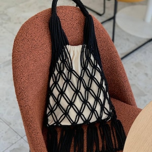 Peut inclure: Un sac fourre-tout en macramé noir et crème avec un motif en filet tissé et des franges. Le sac a un intérieur crème et des anses noires. Le sac est posé sur une chaise marron.