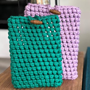 Peut inclure: Deux étuis de téléphone crochetés, un vert d'eau et un lavande, sont présentés côte à côte. Chaque étui est doté d'une fermeture à bouton en bois. Le texte "Crochet Pattern Only" est visible en bas de l'image. Les étuis sont en fil épais et texturé.