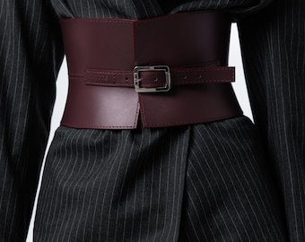 Op de afbeelding: Een bordeauxrode leren corsetriem met een zilveren gesp, gedragen over een donkergrijs krijtstreepjespak. De riem heeft een breed ontwerp en een gespsluiting.