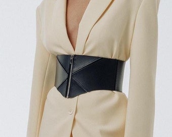 May include: A woman wearing a cream-colored blazer with a black corset belt. The blazer has long sleeves and a V-neckline. The corset belt is wide and has a geometric design.