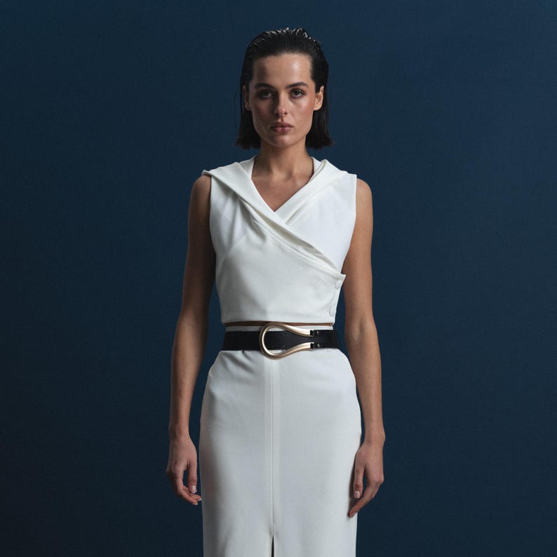 May include: A sleeveless white top and matching skirt with a high waist, paired with a black belt featuring a gold-colored buckle. The outfit is set against a dark blue background, creating a contrast.