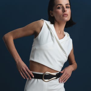 May include: A sleeveless white top with a draped neckline and a black belt with a gold-colored buckle. The outfit is paired with white pants. The background is a solid dark blue.