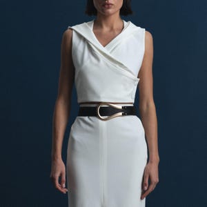 May include: A sleeveless white top with a draped neckline and a matching white skirt. A black belt with a gold-colored buckle cinches the waist. The outfit is set against a dark blue background.