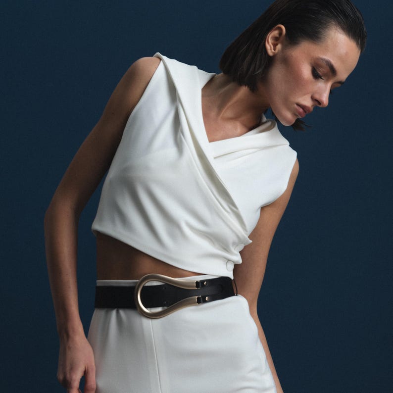 May include: A white sleeveless top with a draped neckline and a black belt with a gold-colored buckle. The model is wearing white pants. The background is a solid dark blue.