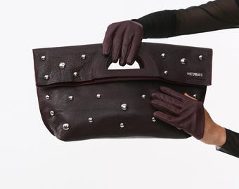 May include: A dark burgundy leather clutch with silver studs and a handle. The clutch is held by a person wearing matching burgundy gloves. The brand name "NEDBAS" is visible on the front.