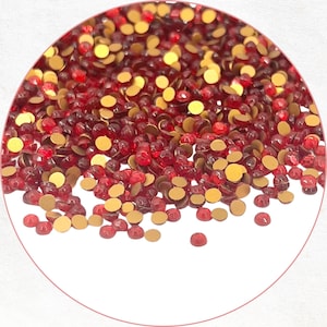 May include: A close-up of a collection of red and gold rhinestones. The red rhinestones are faceted and appear to be made of glass or a similar material. The gold rhinestones are flat and circular. The rhinestones are scattered across a white surface.