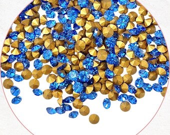 May include: A close-up of a collection of blue and gold rhinestones. The rhinestones are scattered across a white background, with a pink circle border. The blue rhinestones are a vibrant, sparkling color, while the gold rhinestones have a metallic sheen.