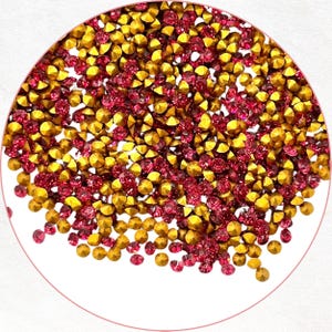 以下が含まれることがあります： 金とルビーレッドの色合いの、小さくファセットカットされたラインストーンの円形配置。宝石は密集して詰め込まれ、きらびやかで質感のある表面を作り出しています。ラインストーンは白い背景に配置されています。