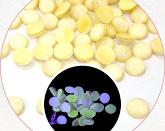 May include: A scattering of pale yellow, circular, flat-backed beads on a white surface. A smaller inset circle displays similar beads glowing with blue and green tones against a black backdrop. These beads are likely for crafting projects.