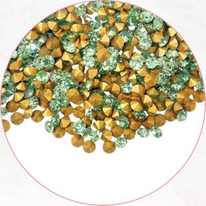 May include: A circular arrangement of gold and pale green rhinestones. The gemstones have a faceted design, catching the light. The image is set against a white background, highlighting the contrast. Ideal for craft projects or embellishments.