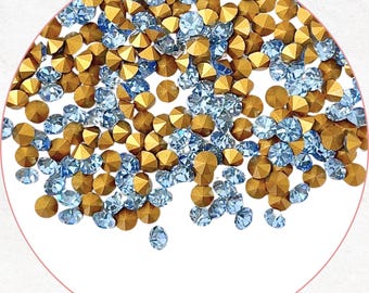 May include: A close-up of a collection of gold and light blue rhinestones. The gold rhinestones have a faceted, geometric shape, while the light blue rhinestones are round. The rhinestones are scattered across a white surface.