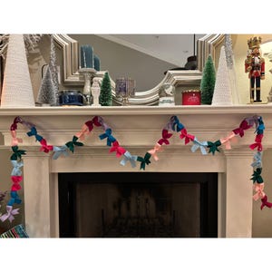 Puede incluir: Una colorida guirnalda de lazos en tonos rosa, azul, verde y morado cuelga frente a una chimenea. La guirnalda está colgada sobre la repisa, añadiendo un toque festivo a la habitación. Los lazos están espaciados uniformemente y crean una exhibición alegre.
