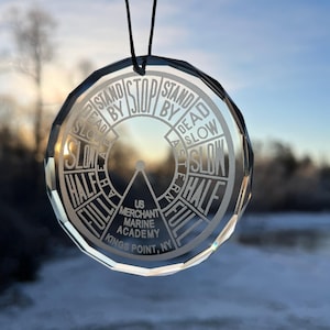 May include: A clear glass ornament with a compass-like design. It features the words "Stand by Stop by Dead Slow Astern Full Half" and "US Merchant Marine Academy Kings Point, NY".