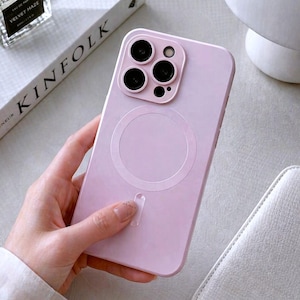 May include: A pale purple phone case featuring a circular design on the back. The case has a camera cutout with three black lenses and a small lens. The case is shown with a white book and a perfume bottle.
